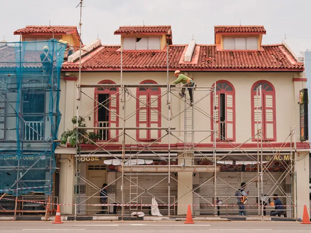 Workers on scaffolding renovating a historic building facade with red roof and tall windows. Renovering af erhvervsejendomme: planlægning, krav og budget